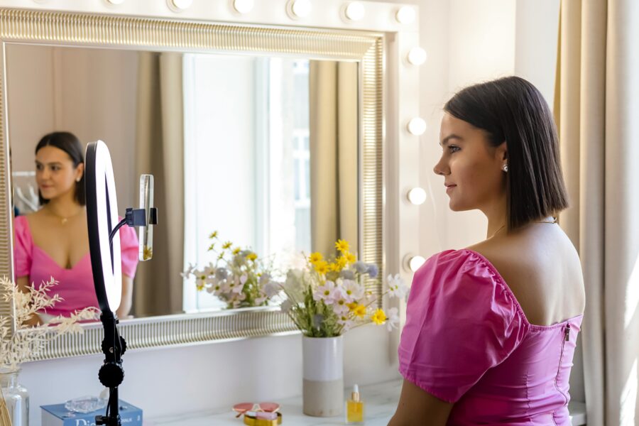 a woman in a pink dress in front of a dressing table mirror with a smartphone and ring light