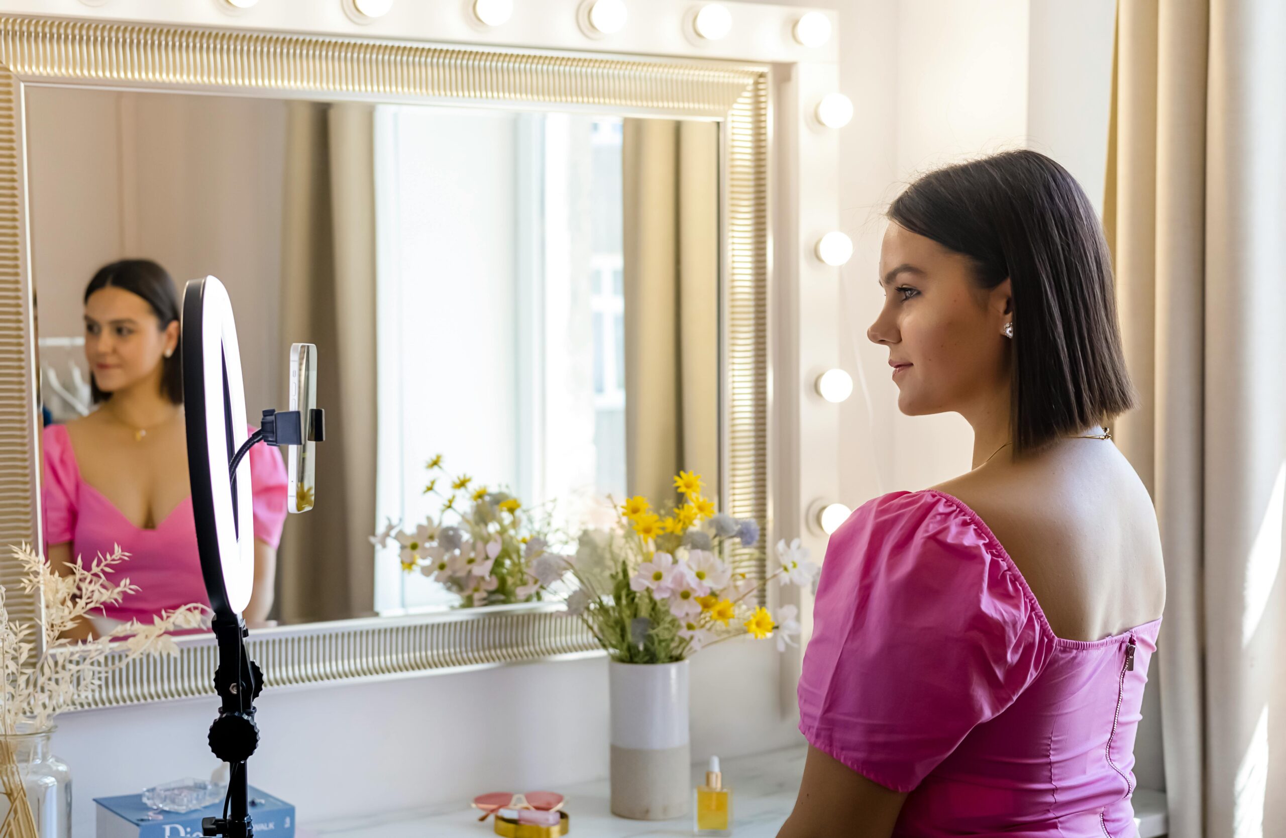 a woman in a pink dress in front of a dressing table mirror with a smartphone and ring light