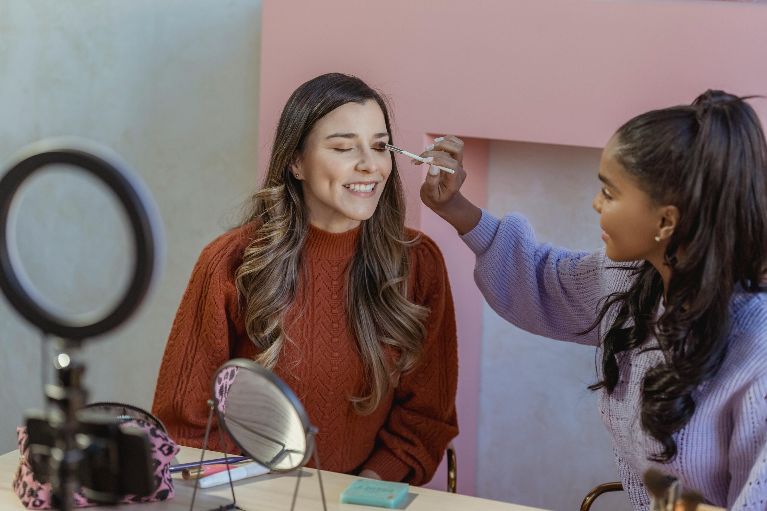 a woman putting makeup on another woman as they film in front of a ring light